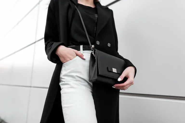 Young stylish woman in a fashionable long black coat in vintage white jeans in a black trendy t-shirt with a leather stylish black handbag is standing outdoors.