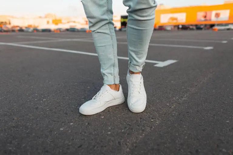 fashionable modern young man in stylish blue jeans in a trendy leather seasonal white sneakers stands in a parking lot in the city
